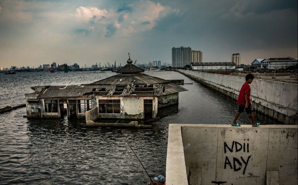 A little boy in Jakarta, walking by a house that has been submerged in water due to rising sea levels
